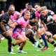 In the image, a dynamic moment from a rugby match is captured, showcasing a player in a vibrant pink kit, driving forward with the ball against the opposition. Surrounding him are players from the opposing team, dressed in black kits, who are intensely focused on halting his advance. The intensity and focus of the players are palpable, highlighting the physicality and fast-paced action typical of a high-stakes rugby game. This scene perfectly illustrates the competitive spirit and athleticism inherent in the sport.