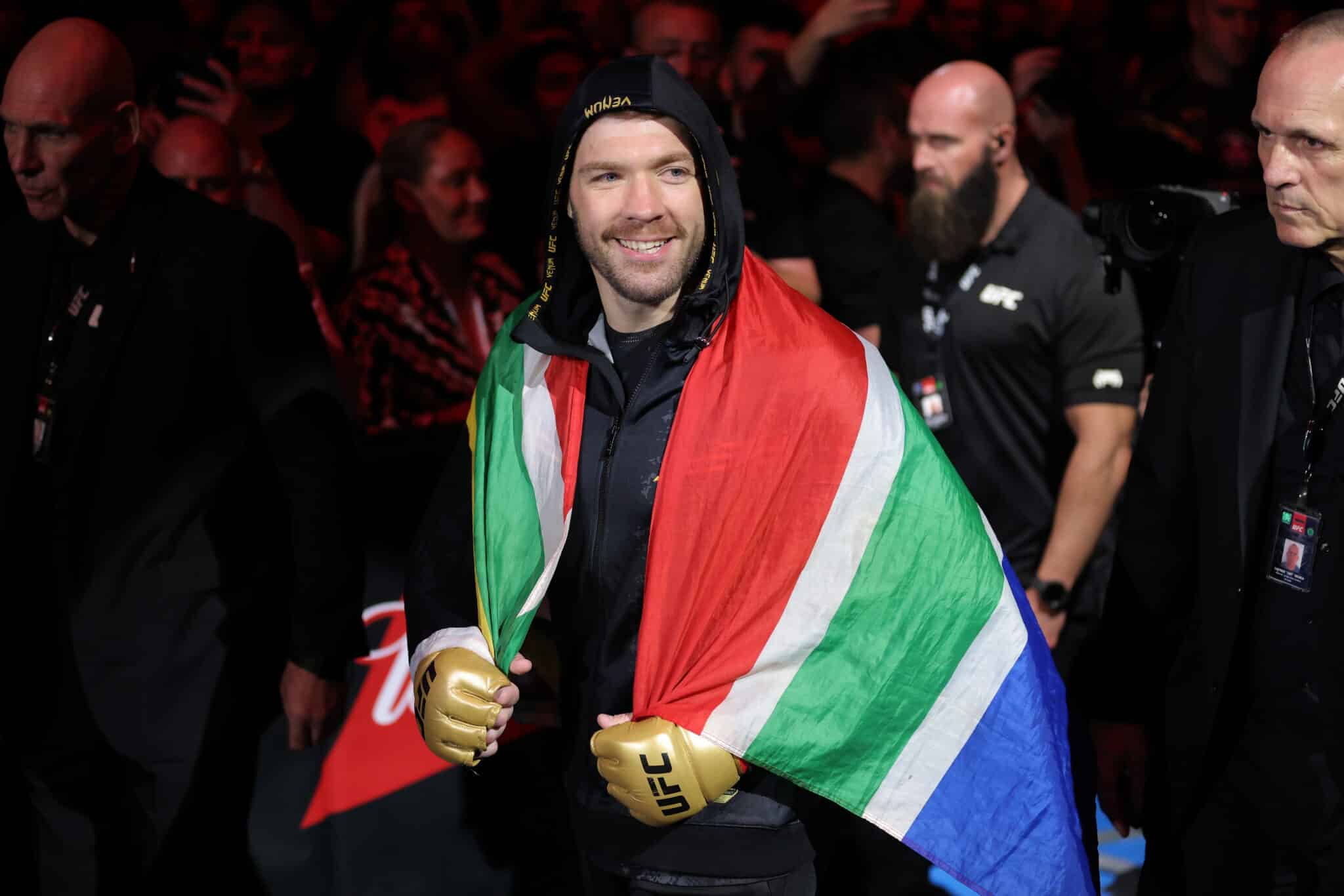 A South African MMA fighter draped in the national flag, wearing gold gloves, smiles as he makes his entrance at a UFC event, surrounded by his team and security.