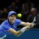 This image captures a tennis player in intense action during a match, dressed in a bright blue shirt with a national flag emblem, suggesting he's playing in a significant international event. He is focused on a low, forceful backhand stroke, showing excellent form and concentration. The blurred background emphasizes the speed and dynamism of the game, highlighting the player's athletic prowess and the high stakes of the competition.