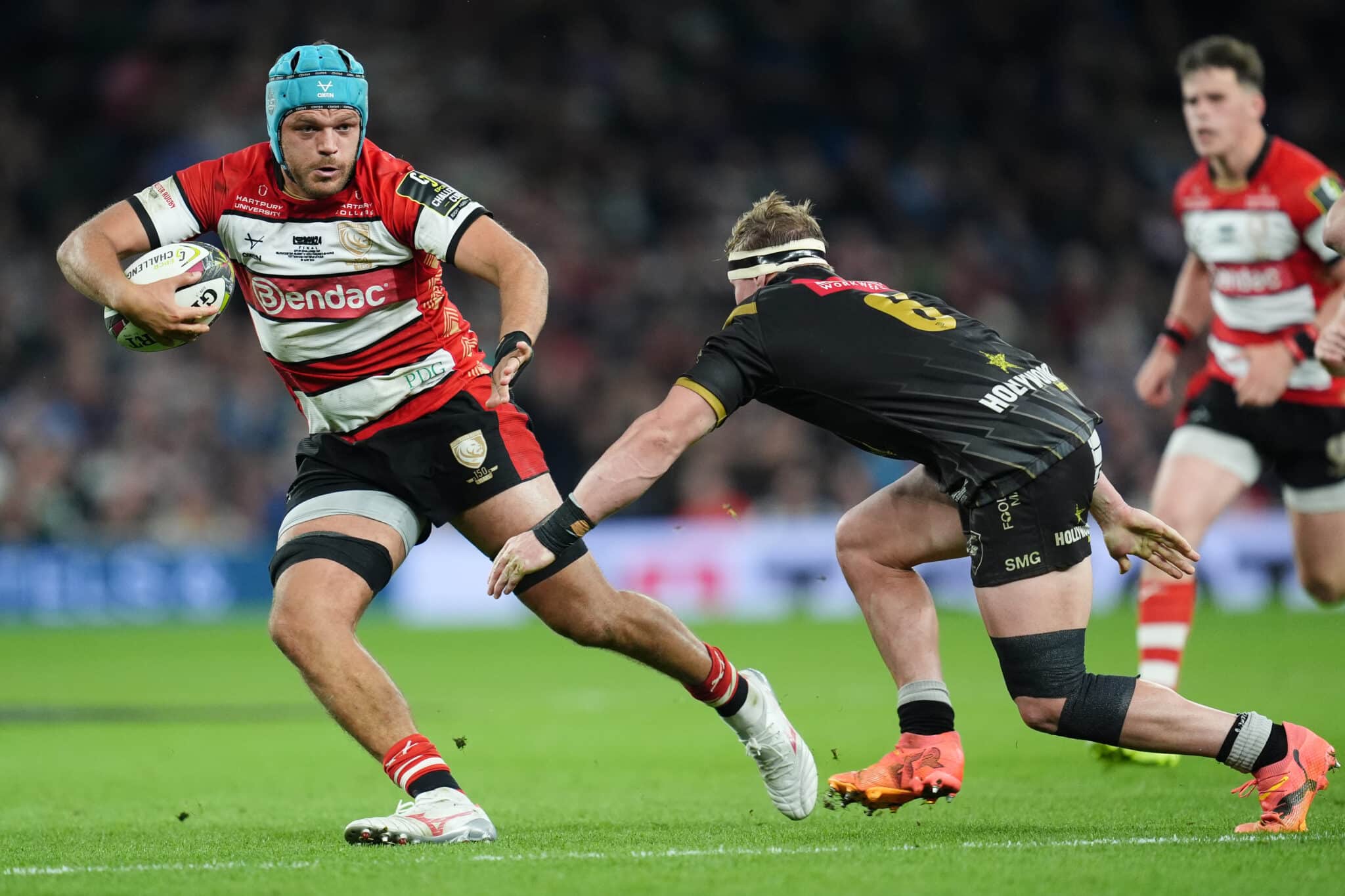 A rugby player in a red and white jersey, identified as a member of the Gloucester Rugby team, runs powerfully with the ball, fending off a tackle from an opposing Exeter Chiefs player in a black and gold uniform during a match.