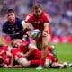 A rugby player in a red jersey from Toulouse deftly passes the ball during a dynamic play, as opponents from Leinster in navy blue attempt to tackle him, showcasing intense action on the rugby field.