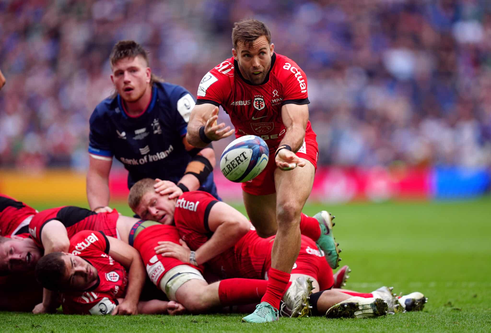 A rugby player in a red jersey from Toulouse deftly passes the ball during a dynamic play, as opponents from Leinster in navy blue attempt to tackle him, showcasing intense action on the rugby field.