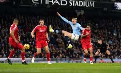 The image captures a dramatic moment from a football match, possibly between Manchester City and a team in red, judging by the uniforms. A player in Manchester City's light blue kit performs an acrobatic kick to play the ball, surrounded by opposing players in red. This action-packed scene occurs under the floodlights of Etihad Stadium, indicating an evening game filled with high energy and athleticism. Such images are emblematic of the intense competition in top-tier football matches, where every move can significantly impact the game's outcome.