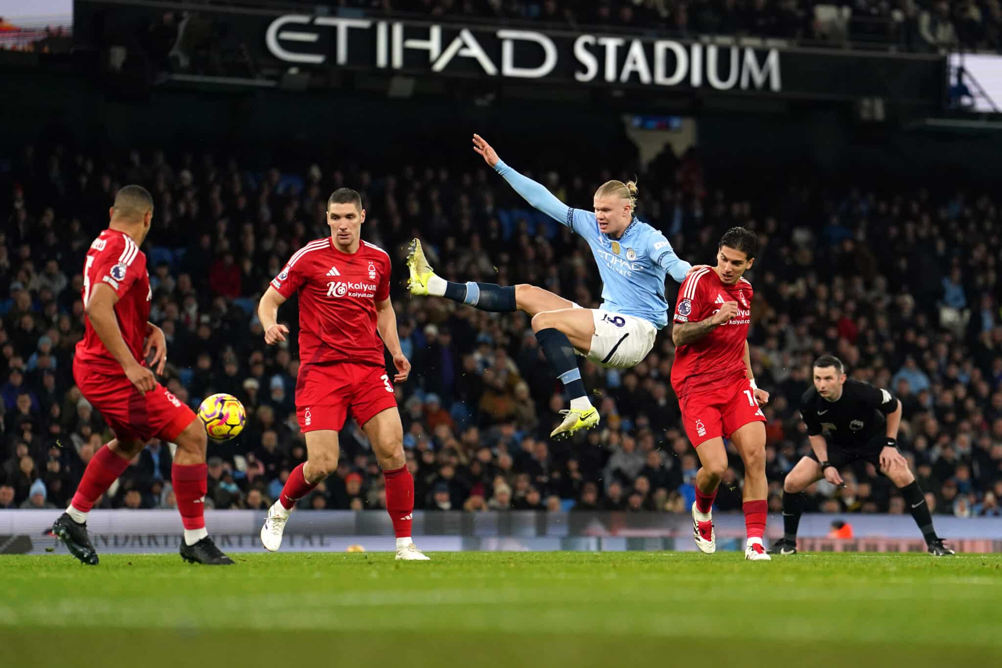 The image captures a dramatic moment from a football match, possibly between Manchester City and a team in red, judging by the uniforms. A player in Manchester City's light blue kit performs an acrobatic kick to play the ball, surrounded by opposing players in red. This action-packed scene occurs under the floodlights of Etihad Stadium, indicating an evening game filled with high energy and athleticism. Such images are emblematic of the intense competition in top-tier football matches, where every move can significantly impact the game's outcome.