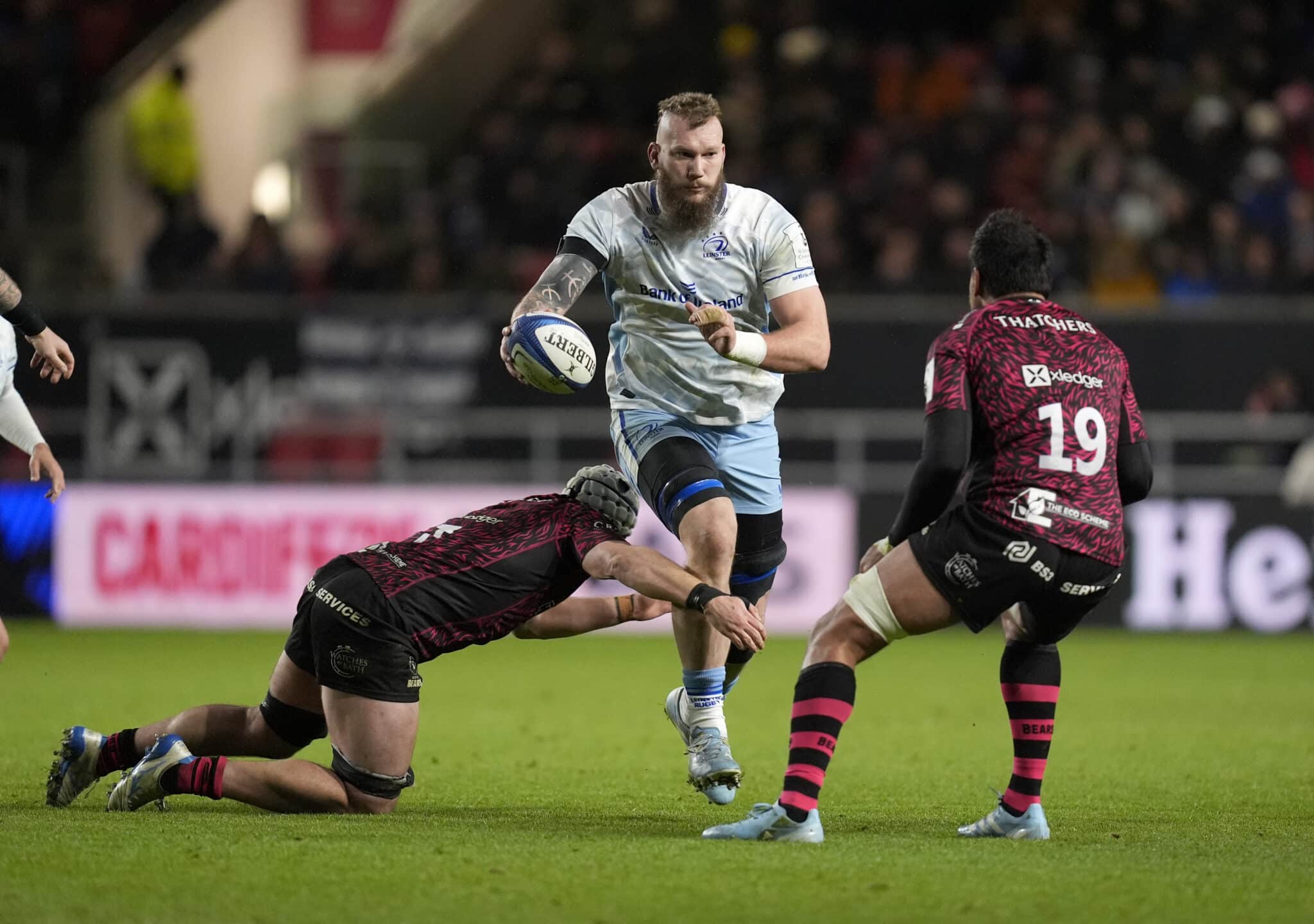 This image captures an intense moment from a rugby match. The player in the white jersey, showcasing determination, is running with the ball, while two opponents in black and pink-striped jerseys attempt to tackle him. The scene is a vivid display of the physicality and dynamic action typical of rugby, highlighting the players' focus and athleticism during the game.