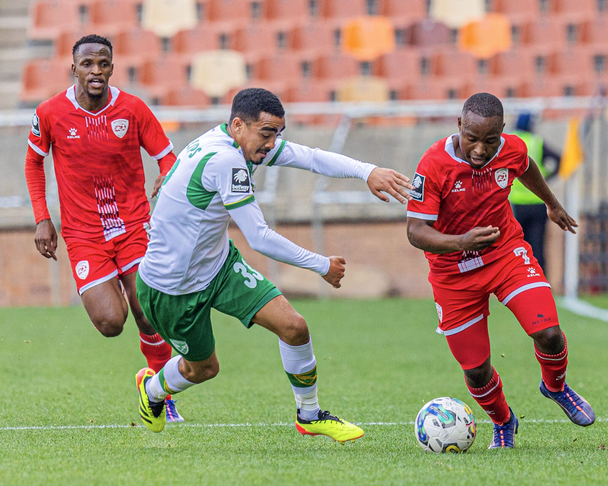 The image captures an intense moment from a football match, featuring three players in a tight contest for the ball. Two players, dressed in red uniforms, are from the same team, while the third player, in a green and white uniform, represents the opposition. The player in green is actively trying to maneuver around his opponents, displaying agility and focus. The backdrop shows a stadium, suggesting the scene is during a competitive league game. The athletes' expressions and body postures indicate the high stakes and fast-paced nature of the match.