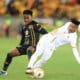 Two soccer players are competing fiercely for the ball during a match. One player, dressed in a black and gold uniform, is maneuvering to control the ball while the other, in a white kit with gold accents, is attempting a tackle or block.