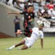 A dynamic action shot from a soccer match showing a player from Orlando Pirates in black and red gear jumping to head the ball, while a player from a team in white with a gold emblem attempts a tackle on the ground, with spectators and a cameraman in the background.