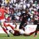 The image captures an intense moment from a CAF Champions League match, showing a clash between Orlando Pirates, in their black and red uniforms, and Al-Ahly, with players in red. An Orlando Pirates player is seen executing a dynamic slide tackle or falling to the ground as he attempts to intercept the ball from an Al-Ahly player who is dribbling forward.