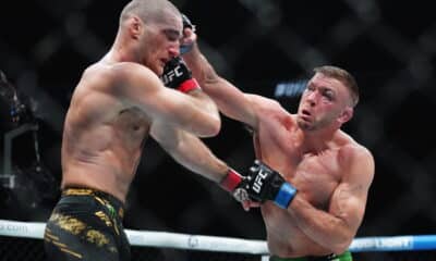 In the image, two UFC fighters are intensely engaged in combat during a match. One fighter, wearing black and gold shorts, is landing a powerful punch on his opponent, who is in green shorts. The impact of the punch is visibly jarring, demonstrating the high stakes and fierce competition in UFC fights. Both athletes display concentration and physical prowess, capturing the dramatic and brutal essence of mixed martial arts competitions.